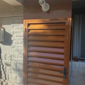 Brown metal door with horizontal louvers mounted in a stone exterior wall; visible door handle and hinge on the left side, sunlight casting shadows on the surface.