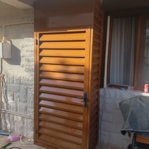 Brown metal louvered vent/door standing outside beside a glass door on a stone wall, with tools and a grill nearby.